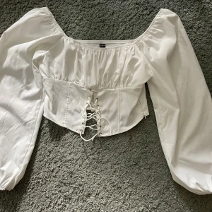 Vit korsettopp med puffärmar och snörning - Superfin vit korsettopp  med bred offshoulder-ringning, puffiga långärmar och snygg snörning framtill. Toppen är croppad och har dragkedja i sidan för enkel påklädning. Perfekt för dig som vill sticka ut med en trendig look.