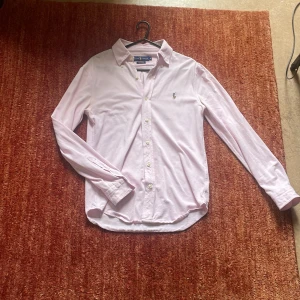 Ljusrosa skjorta från Ralph Lauren  - Säljer en klassisk ljusrosa skjorta från Ralph Lauren i storlek S. Skjortan har lång ärm, knappar framtill och ikoniska broderade loggan på bröstet. Tillverkad i bomull med en snygg oxfordstruktur. Perfekt för dig som gillar stilrena och tidlösa plagg. Bra skick! Kom med prisförslag.