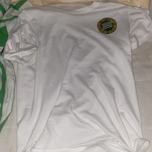 Vit Hammarby Södermalm t-shirt - Vit t-shirt med grönt Hammarby Södermalm-tryck på ryggen och klubbmärke på bröstet. Klassisk passform och rund hals. Perfekt för dig som vill visa ditt stöd för Hammarby. Skön och enkel stil som funkar året runt.