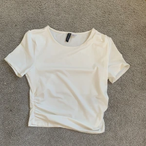 Vit kortärmad topp från Divided - En stilren vit topp från Divided by H&M i storlek M. Modellen har korta ärmar, rund halsringning och lätt rynkade detaljer i sidorna.