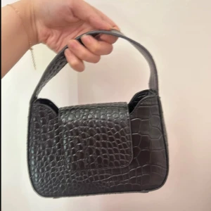 Svart axelväska i krokopräglat skinn - Snygg svart axelväska med croco-mönster och kort rem. Väskan har en stilren fyrkantig form och stängs med lock. Perfekt för att lyfta vilken outfit som helst med en trendig touch.