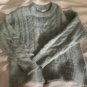 Grå stickad tröja från Gina Tricot - Mysig grå stickad tröja från Gina Tricot med grovt kabelstickat mönster. Tröjan har rund halsringning, ribbade muddar och lång ärm. Perfekt för lager-på-lager och chill höst- och vinterstil. Storleken är S men känns mer som en M💓