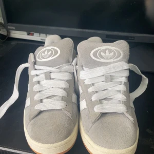 Adidas Campus grå sneakers mocka - Snygga Adidas Campus sneakers i ljusgrå mocka med vita detaljer och klassiska tre ränder på sidan. Skorna har vit sula med gummiyttersula och vita skosnören. Perfekta för en clean och stilren look.