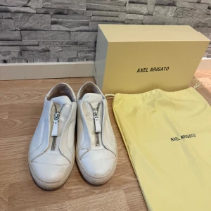 Vita sneakers Axel Arigato med dragkedja - Stilrena vita sneakers från Axel Arigato i skinn med unik dragkedja framtill istället för klassisk snörning. Låg modell med rund tå och diskret logga på sidan. Kommer med originalkartong och dustbag. Perfekta för dig som gillar minimalistisk och modern stil.