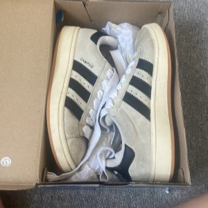 Adidas Campus sneakers i beige mocka - Snygga Adidas Campus sneakers i beige mocka med klassiska svarta ränder på sidorna och vit sula med gummidetalj. Skorna har vit snörning och en stilren, retro vibe som passar perfekt till streetwear. Ikonisk Campus-logga på sidan. Har tyvärr inte kvar orginalkartongen men har lagt de i en annan kartong