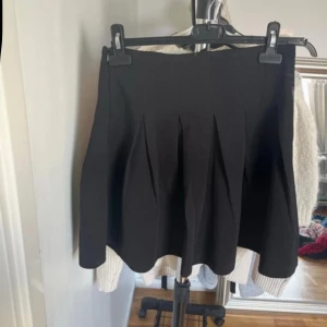 Svart plisserad kortkjol - Snygg svart kortkjol med plisserade veck som ger en cool och trendig look. Kjolen har en klassisk A-linjeform och passar perfekt till både sneakers och boots. Enkel att matcha med olika toppar för en personlig stil.