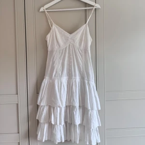 Vit volangklänning - Superfin vit klänning med smala axelband, v-ringning och dekorativa knappar framtill. Klänningen har volanger i kjolen, spetsdetaljer och broderade partier. Dragkedja i sidan för enkel påklädning. Perfekt för ljusa dagar och studentfirande!