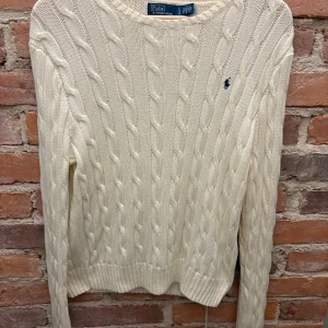 Vit kabelstickad tröja Polo Ralph Lauren - Kabelstickad vit tröja från Polo Ralph Lauren med klassisk rund hals och broderad marinblå logga på bröstet. Tröjan har lång ärm och ribbade muddar, perfekt för lager-på-lager och snygg till jeans😉
