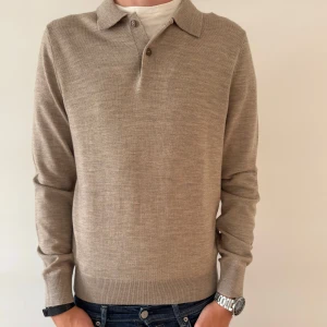 MORRIS MERINOULL TRÖJA - Snygg beige pikétröja från Morris i mjuk merinoull. Klassisk krage, knäppning med två knappar och lång ärm. Perfekt för dig som gillar stilrena och tidlösa plagg. Tröjan är tunnstickad och har ribbade muddar vid ärmslut och nederkant.