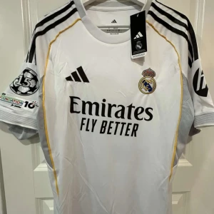 Real Madrid Mbappé 10 tröja - Snygg vit Real Madrid fotbollströja från med svarta detaljer och gula inslag. Tryck med Mbappé och nummer 10 på ryggen. Klassisk klubbmärke på bröstet och Champions League-märke på ärmen. Tillverkad i lätt och ventilerande material. Oanvänd och kommer med taggar 