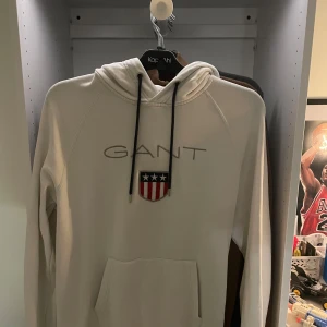 Vit hoodie från GANT  - Snygg vit hoodie från GANT med broderad logga och sköld på bröstet. Tröjan har huva med svarta snören och en stor magficka. Perfekt för dig som gillar klassisk och stilren streetwear. Storlek L. Aldrig använd