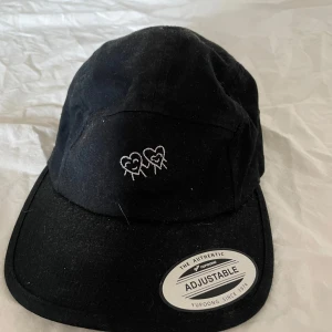 Svart keps från Yupoong med brodyr - Svart keps från Yupoong i 100% bomull med justerbar passform. Framtill finns två broderade hjärtan och på sidan står det 'Love u <3' i vit brodyr. Klassisk och stilren modell som passar till det mesta.