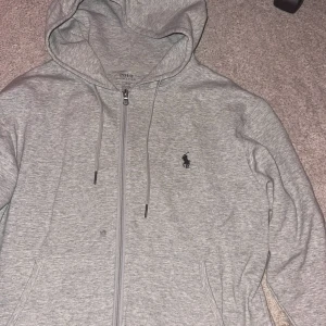 Grå hoodie från Polo Ralph Lauren - Snygg ljusgrå hoodie från Polo Ralph Lauren med dragkedja och klassisk logga på bröstet. Tröjan har huva med snörning och två fickor framtill. Perfekt för en chill och stilren look. Byten funkar kan gå ner i pris vid snabb affär