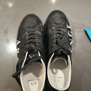 Svarta sneakers från Armani Exchange - Svarta sneakers från Armani Exchange med vit logga längs sidan. Skorna har snörning, vit insida och en robust platt sula. Tillverkade i syntetmaterial med klassisk rund tå. Perfekta för dig som gillar stilrena och trendiga sneakers.
