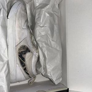 Nike Book 1 sneakers vit/brun  - Nike Book 1 sneakers i vitt och ljusbrunt med coolt ormmönstrat swoosh på sidan. Skorna har snörning, platt sula och är tillverkade i en mix av skinn och syntet. Perfekta för dig som gillar unika detaljer och streetstyle.