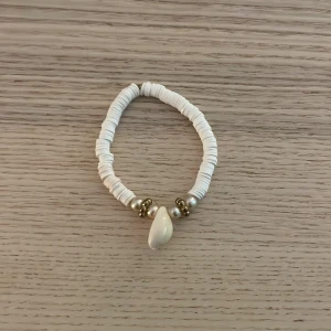 Vitt armband med snäcka och pärlor - Superfint armband med vita platta pärlor, guldfärgade detaljer och en snygg snäcka i mitten. Perfekt accessoar för en somrig vibe och strandkänsla. Stretchig modell som passar de flesta handleder.