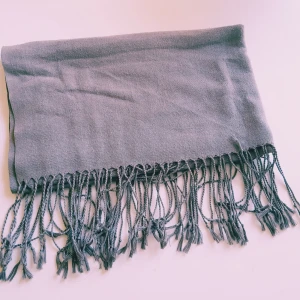 Grå halsduk med fransar - En stilren grå halsduk med långa tvinnade fransar i ändarna. Halsduken är mjuk och har en enkel, klassisk design som passar perfekt till jackor och kappor. Perfekt accessoar för kyliga dagar. Har 2 likadana.  Helt skick dock noppror