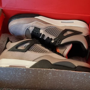Nike Air Jordan 4 Taupe Haze sneakers - Nike Air Jordan 4 Taupe Haze med grå mocka, svarta detaljer och meshpaneler. Snygg kontrast med röd Jumpman-logga på plös och sula. Klassisk siluett med svart snörning och vit mellansula. Perfekta för dig som vill sticka ut med stilrena och trendiga sneakers.