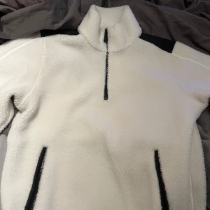 Vit fleece tröja från Sail Racing storlek S - Vit teddyjacka från Sail Racing i storlek S. Jackan har svart panel över axlarna, halv dragkedja framtill och två fickor med svarta dragkedjor. Mjuk och fluffig känsla, perfekt för kyliga dagar. Enkel och stilren design med hög krage.