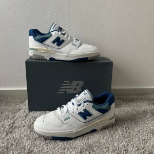 New Balance 550 vita och blå sneakers - Snygga New Balance 550 sneakers i vitt och marinblått med detaljer i ljusblått. Skorna har klassisk retrodesign, snörning och platt sula. Tillverkade i skinn och syntet, perfekta för dig som gillar streetstyle och sportig look.