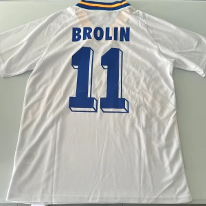 Sverige Brolin 11 fotbollströja Adidas - Klassisk vit Sverige fotbollströja från Adidas med blå och gula detaljer, nummer 11 och Brolin på ryggen. Tröjan har korta ärmar, V-ringad krage med blå, gul och röd kant samt broderat landslagsmärke på bröstet. Perfekt för dig som älskar retro och landslagskänsla. Endast handtvätt gäller. Replika