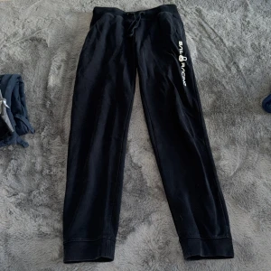 Svarta mjukisbyxor Sail Racing - Svarta mjukisbyxor från Sail Racing med vit logga på ena benet. Byxorna har snörning i midjan och muddar vid bensluten. Perfekta för chill dagar eller när du vill vara bekväm men ändå se snygg ut.