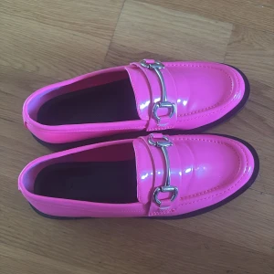 Glansiga rosa loafers med metallspänne - Snygga loafers i en stark rosa färg med glansig finish och silverfärgat metallspänne över vristen. Skorna har en klassisk rund tå och platt sula med låg klack. Perfekta för dig som vill sticka ut med färg och stil.