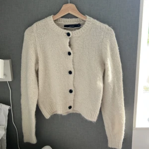 Beige stickad kofta från Vero Moda - Supermysig beige stickad kofta från Vero Moda med svarta knappar framtill. Koftan har långärmad passform, rund halsringning och ribbade muddar. Perfekt för lager-på-lager och chill höst- och vinterstil.