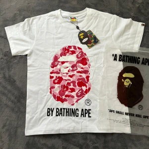 Vit BAPE t-shirt med rosa camo-tryck - Vit t-shirt från BAPE med stort ikoniskt apansikte i rosa och röd camo på bröstet. Klassisk rund hals och korta ärmar. Tryckt text 'BY BATHING APE' nedanför motivet. Perfekt statement-plagg för streetwear.