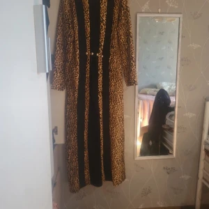 Långklänning med leopardmönster och svarta paneler - Cool långklänning med leopardmönster och svarta vertikala paneler. Klänningen har långa ärmar och markerad midja med dekorativa knappar framtill. Perfekt för dig som vill sticka ut med en djärv look.