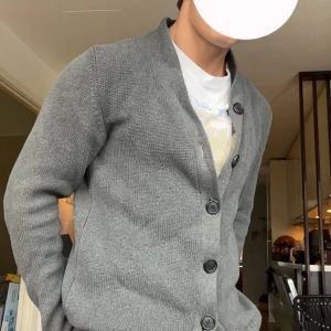 Grå kofta med stora knappar H&M - Snygg grå kofta från H&M med stora svarta knappar och ribbade muddar. Koftan är stickad och har en klassisk krage som ger en chill och stilren look. Perfekt att slänga på sig över en t-shirt.