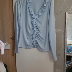 Ljusblå långärmad topp med volang - Supersöt ljusblå långärmad topp med djup v-ringning och volangdetalj längs hela framsidan. Mjuk och lätt i materialet, perfekt för dig som gillar romantiska vibes och vill sticka ut lite extra.