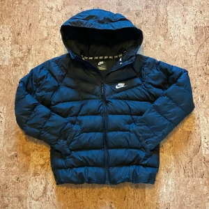 Svart pufferjacka från Nike - Snygg svart pufferjacka från Nike med huva och dragkedja. Jackan har klassisk quiltad design, två fickor och Nike-logga på bröstet. Perfekt för kyliga dagar och enkel att matcha med streetwear. Fodrad för extra värme och har sportig vibe. Storlek 158 som översättts till Xs