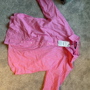 Rosa randig skjorta från Zara XS - Snygg rosa skjorta från Zara i storlek XS med vita ränder och klassisk krage. Skjortan har långa ärmar, knappar framtill och är gjord i mjuk bomull. Perfekt för dig som gillar färg och vill sticka ut lite extra.
