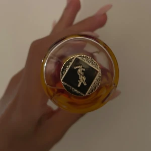 Yves Saint Laurent Manifesto parfym - Säljer en snygg parfymflaska från Yves Saint Laurent, Manifesto. Flaskan är genomskinlig med guldigt lock och en lila/röd detalj i mitten. Parfymen har en lyxig känsla och flaskan har en elegant, kurvig form med YSL-logga i botten.