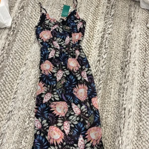 Blommig långklänning från H&M  - Supersnygg långklänning med tropiskt blommönster. Klänningen har smala axelband, markerad midja och flowy passform. Perfekt för sommaren. Helt ny med prislappen på, oanvänd. 
