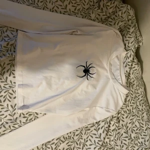 Vit långärmad tröja med spindeltryck - Vit långärmad tröja med ett svart spindeltryck på bröstet. Tröjan har en klassisk rund halsringning och är gjord i mjuk bomull, perfekt för dig som gillar unika detaljer och vill sticka ut lite extra.