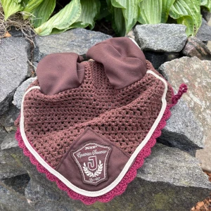 Brun ridmössa från Jacson - Snygg brun ridmössa från Jacson aldrig använt med virkad design och tyg över öronen. Märke framtill och vinröd kant ger en extra touch. Perfekt för ridning och skyddar hästens öron mot insekter och ljud.