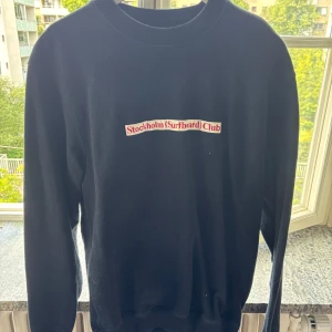 Svart sweatshirt från Stockholm Surfboard Club - Svart sweatshirt från Stockholm Surfboard Club med röd och vit logga på bröstet. Tillverkad i 100% ekologisk bomull, har rund hals och långa ärmar. Enkel och clean design som passar perfekt till jeans eller shorts.