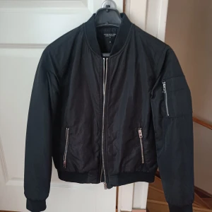 Svart bomberjacka med dragkedjor - Snygg svart bomberjacka.. Jackan har flera dragkedjor, bland annat på fickorna och ärmen, samt ribbade muddar vid ärmslut och midja. Perfekt för dig som gillar en stilren och urban look.