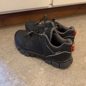 Svarta sneakers med orange detaljer - Svarta sneakers med robust sula och orange detaljer på hälen. Skorna har snörning och är tillverkade i syntetmaterial med meshfoder. Vattenavvisande och perfekta för vardagsbruk eller promenader.