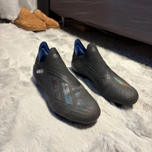 Adidas X svarta fotbollsskor - Snygga Adidas X fotbollsskor i svart med blå detaljer och diskreta regnbågsskimrande ränder på sidan. Skorna har en strömlinjeformad design utan snörning och är tillverkade i syntetmaterial för bästa känsla på planen. Namnen på sidan är borttagna 