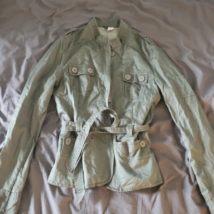 Grön utilityjacka från H&M Divided - Snygg grön utilityjacka från H&M Divided med bälte i midjan, fyra fickor med knappar och coola axeldetaljer. Jackan har lång ärm och stängs med knappar framtill. Perfekt för dig som gillar en avslappnad men ändå stilsäker look.