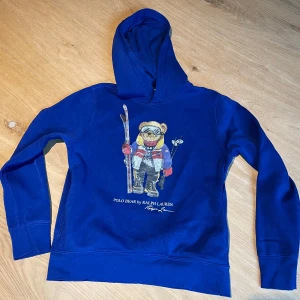 Blå hoodie Polo Bear Ralph Lauren Jr - Blå hoodie från Ralph Lauren med ikoniska Polo Bear-trycket framtill. Tröjan har huva, långa ärmar och är gjord i mjuk bomull. Perfekt för dig som gillar streetstyle och vill ha något som sticker ut med en cool björn i skidoutfit på bröstet.