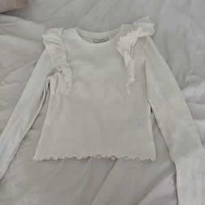 Vit ribbad tröja med volang från Kappahl - Supersöt vit långärmad tröja från Kappahl med ribbad struktur och volangdetaljer på axlarna. Tröjan har en lätt croppad känsla med vågig kant nertill och är perfekt för dig som gillar en feminin och trendig stil. den är lite smutsig men jag tvättar så klart den innan jag skickar iväg