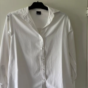 Vit klassisk skjorta från Gina Tricot - Vit skjorta i bomull från Gina Tricot med klassisk krage och långa ärmar. Skjortan har knappar framtill och vid ärmsluten, rak passform och stilren look. Perfekt att styla med jeans eller kjol för en clean vibe.
