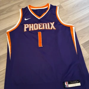 Phoenix Suns Booker basketlinne Nike - Säljer ett lila basketlinne från Nike med Phoenix Suns-tryck och nummer 1, Booker på ryggen. Linnet har orange och vita detaljer längs kanterna och ärmlös design. Perfekt för basketfans som vill sticka ut på planen eller på läktaren.