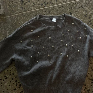 Grå stickad tröja med pärlor XS - Mysig grå stickad tröja i storlek XS med pärldetaljer framtill. Tröjan har rund halsringning och långa ärmar. Perfekt för dig som gillar en enkel men ändå extra look med pärlor som ger en cool touch.