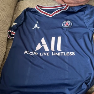 PSG matchtröja blå Jordan x Nike - Snygg blå PSG fotbollströja med Jordan-logga och klubbmärke på bröstet. Klassisk V-ringad krage med röd, vit och blå rand. Kortärmad modell i lätt och ventilerande material, perfekt för träning eller match. Sponsortexten 'Accor Live Limitless' på framsidan.