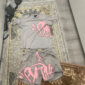 Grå shorts med rosa graffiti från Stayhard - Grå shorts från Stayhard med stor rosa graffiti-print framtill och matchande rosa snörning i midjan. Mjukt material, elastisk midja och bakficka med liten logga. Perfekta för chill dagar eller streetwear-vibe. Dessa funkar utmärkt för folk som vill sticka ut lite men inte se ut som att man försöker för mycket.